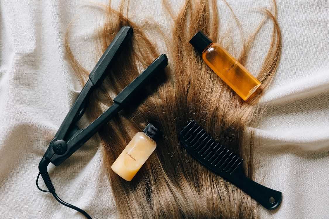 Flat iron, hair oil bottles, comb on spread blonde hair.